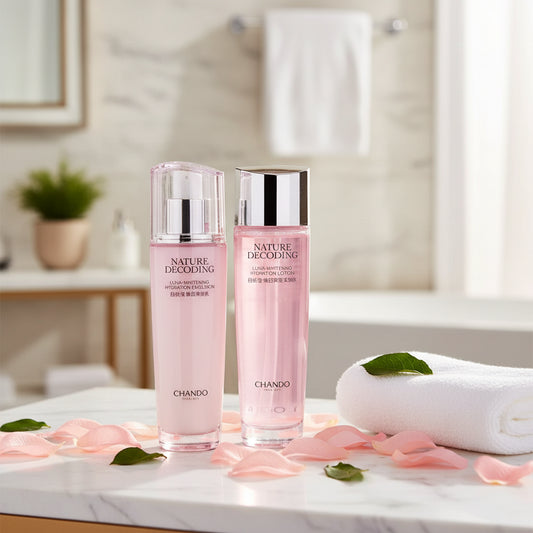 Two pink Chando skincare bottles with silver caps, labeled 'Nature's Decoding', displayed on a marble countertop with pink petals, a white towel, and green leaves in a softly lit bathroom setting.