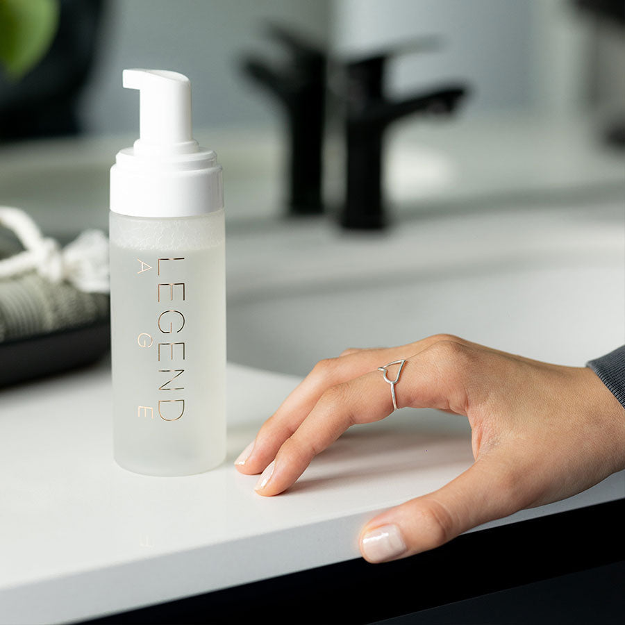 Clear bottle with white pump on a bathroom counter, hand reaching out.