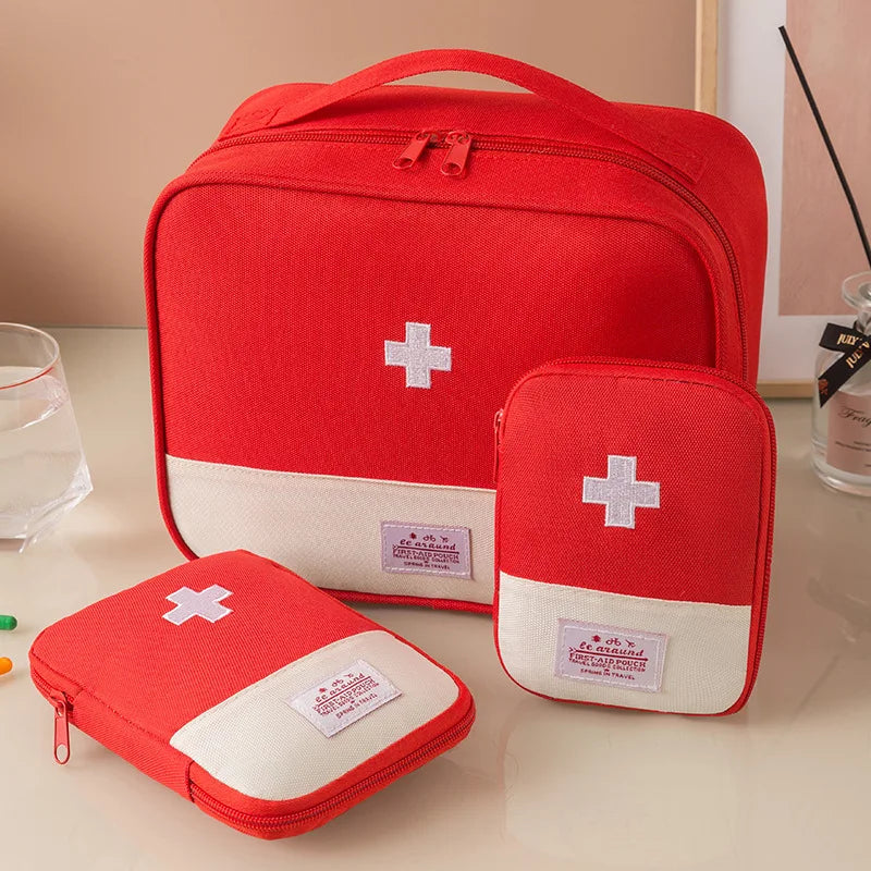 Three red and off-white fabric first aid or travel kit pouches arranged on a tabletop, with a larger rectangular bag behind two smaller zippered pouches. Each pouch has a white cross emblem and a small sewn label.