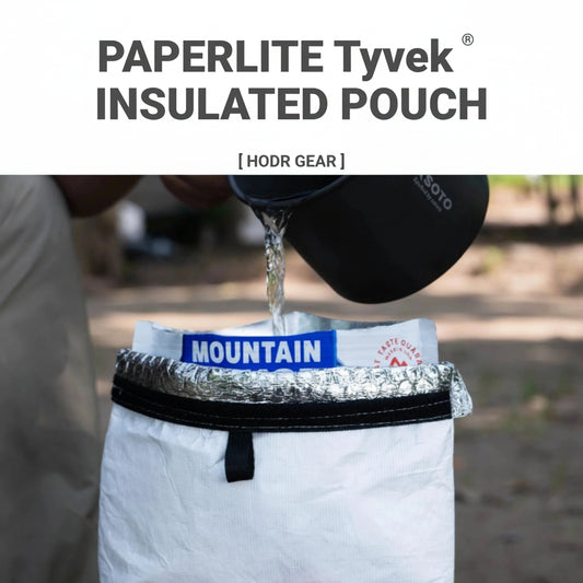A white insulated pouch with black branding and a silver foil-lined opening, shown outdoors with a person pouring hot water from a black mug into it. Large text at the top reads 'PAPERLITE Tyvek INSULATED POUCH' .