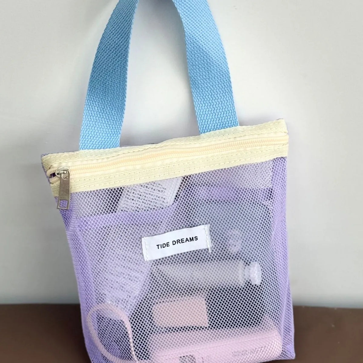 A light purple mesh zippered toiletry bag with light blue handles and a cream-colored zipper trim. A white label on the front reads 'TIDE DREAMS'.