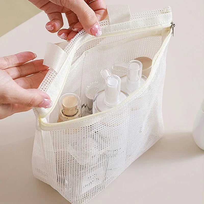 A cream-colored mesh zippered cosmetic bag with two handles, shown being held open by hands to reveal toiletry bottles inside, set against a soft beige background.