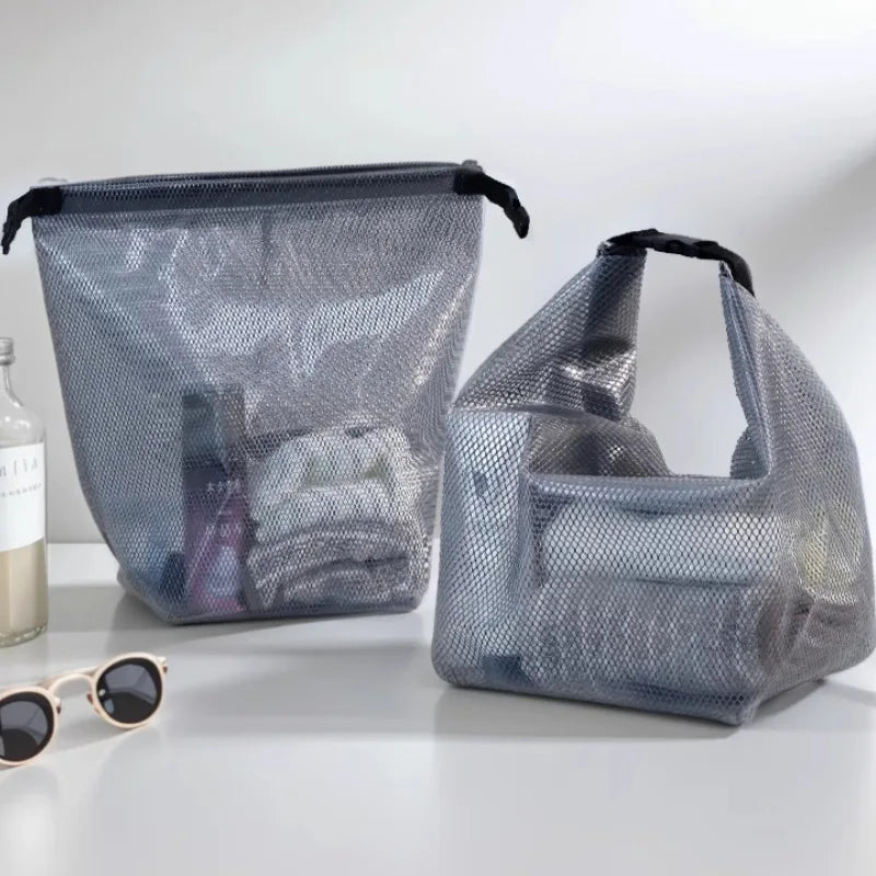 Two gray mesh zippered luggage or travel bags with black handles, shown on a white surface with a bottle and sunglasses nearby.
