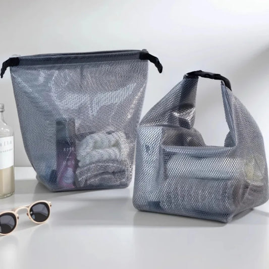 Two gray mesh zippered luggage or travel bags with black handles, shown on a white surface with a bottle and sunglasses nearby.