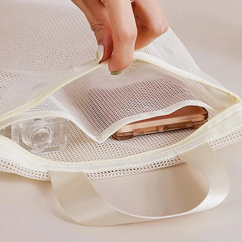 A close-up of a hand opening a cream-colored mesh tote bag, showing a phone and small clear bottle inside the open compartment.