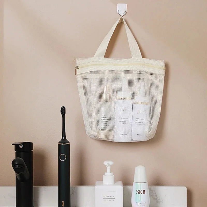 A beige mesh hanging toiletry bag with two handles is suspended from a wall hook above a bathroom counter. The bag contains several white skincare bottles, and below it are a black electric toothbrush and other bathroom items against a warm beige wall.