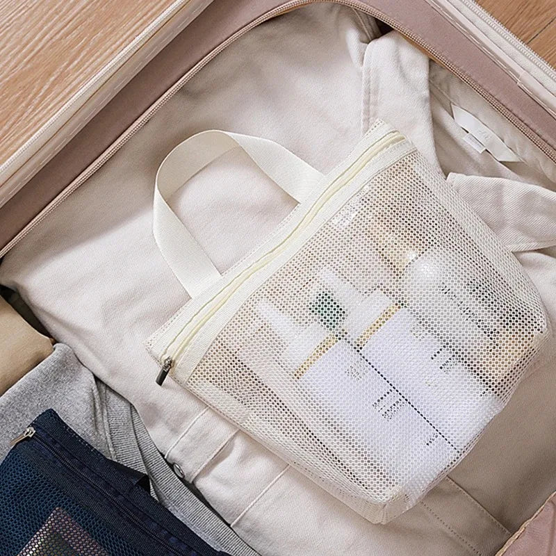 A white mesh zippered toiletry bag with a cream handle, shown inside an open suitcase packed with travel items.