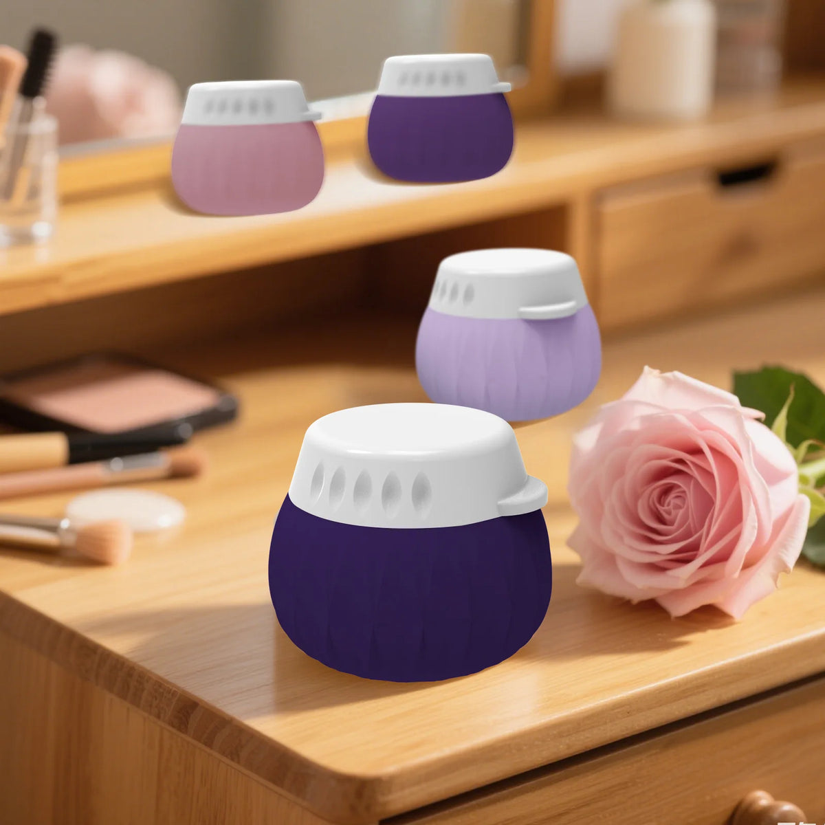 Three small round cosmetic containers in purple and lavender shades with white lids, arranged on a wooden vanity table next to a pink rose, with a blurred mirror reflection in the background.
