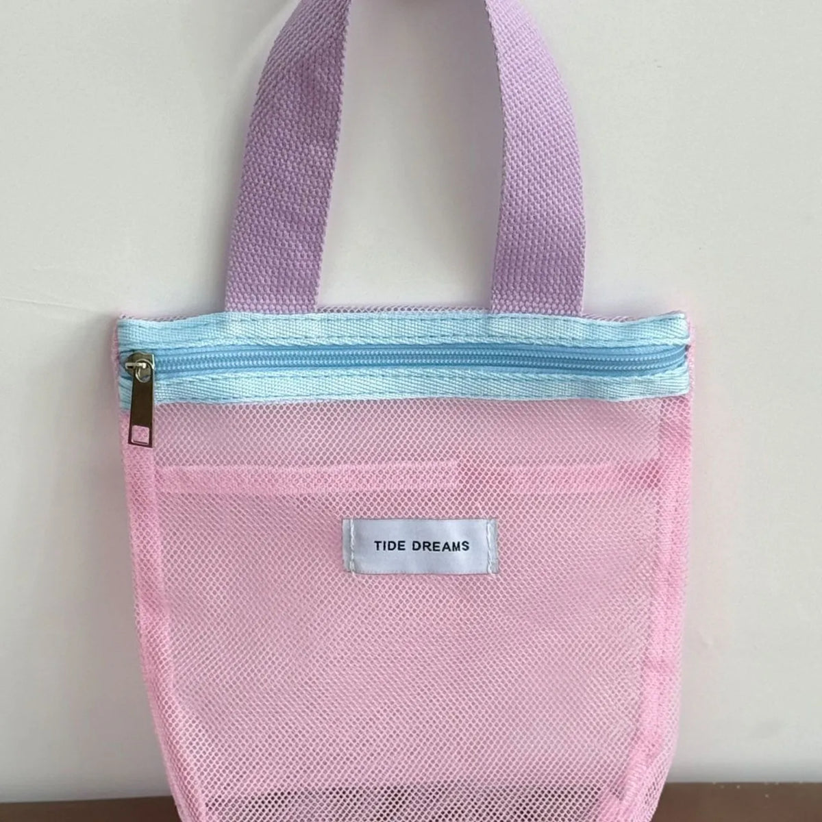 A pink mesh zippered pouch with lavender fabric handles and a light blue zipper trim. A silver label on the front reads 'TIDE DREAMS'.