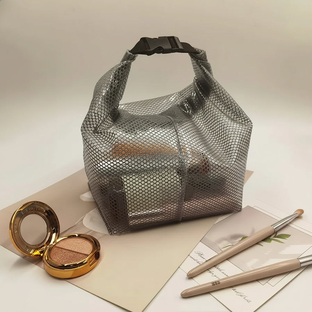 A translucent gray mesh makeup bag with a black top handle, shown on a light beige background with cosmetic props including an open gold compact and makeup brushes.