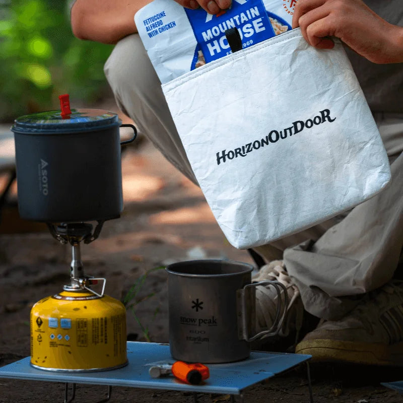 Person with a Horizon Outdoor bag, camping stove, and mug outdoors.