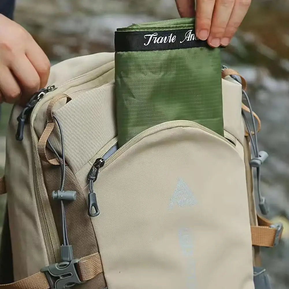 A close-up of a beige backpack with a side pocket holding an olive green rolled item, with a hand lifting the top flap. The flap shows the brand name 'Tivoli Arts' in white script on a black band.