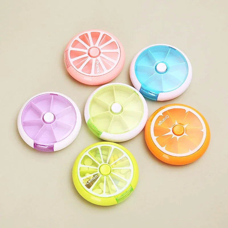 Six small oval compact-style items in assorted citrus and pastel colors, arranged on a plain beige background. The tops resemble sliced citrus fruits, including pink, blue, purple, pale yellow, orange, and lime green.