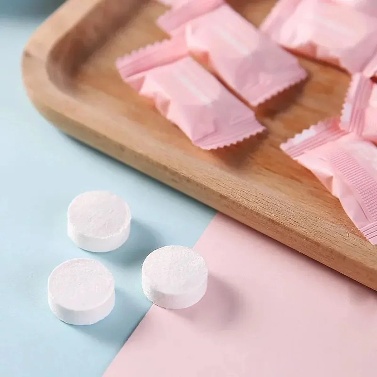 Pink and white chewable tablets on a wooden tray with a light blue background
